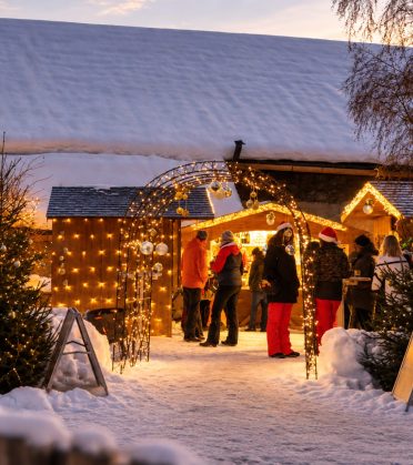 Beleuchtete Holzhütte mit Lichterketten im Schnee, davor Menschen an einem Adventstand in der Abenddämmerung