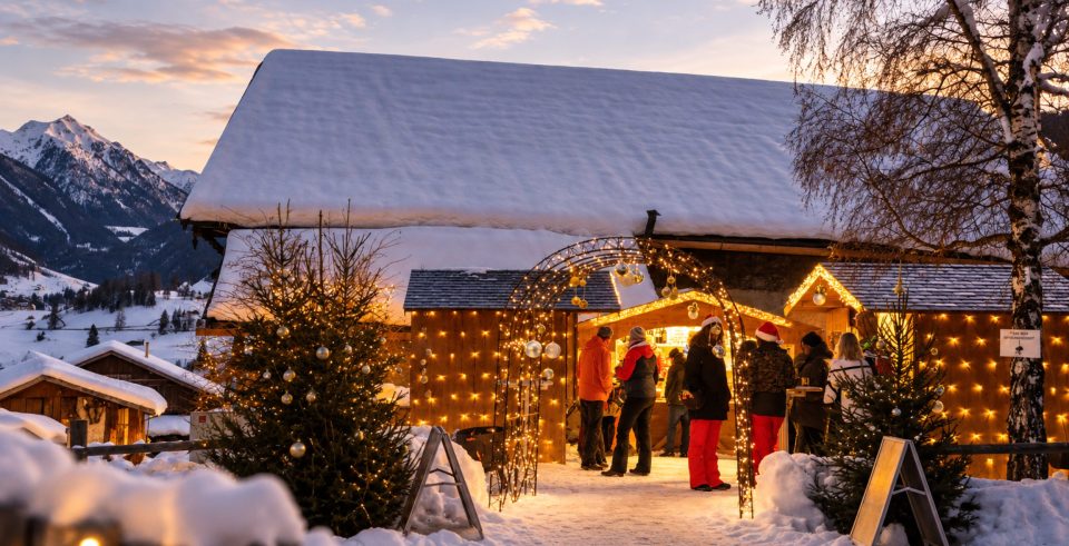 Beleuchtete Holzhütte mit Lichterketten im Schnee, davor Menschen an einem Adventstand in der Abenddämmerung