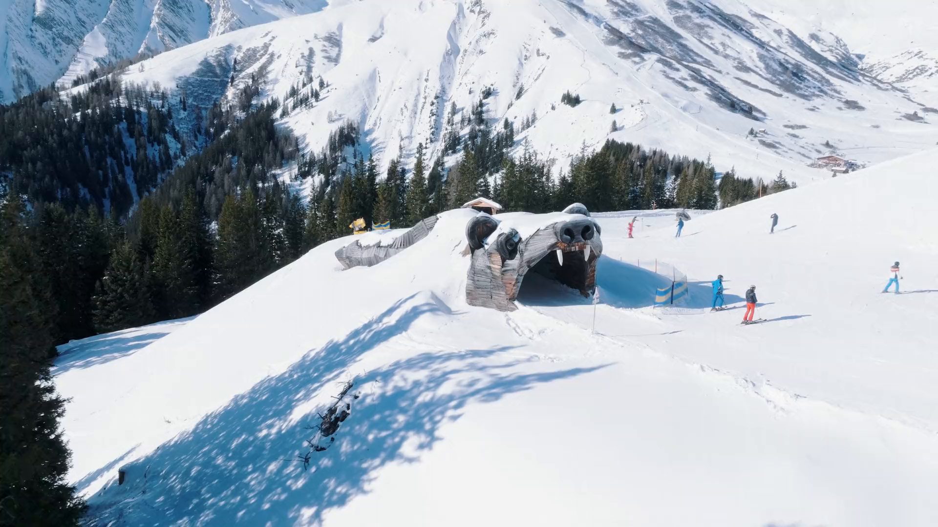 Die Bärenpiste in Serfaus aus der Luft: Skifahrerinnen und Skifahrer fahren durch den hölzernen Bärenkopf, dahinter das verschneite Tal.