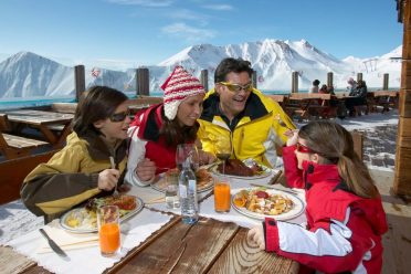 Eine Familie sitzt zur Einkehr an der langen Holztafel auf der Sonnenterrasse, dahinter die verschneiten Berge.