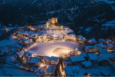 Der beleuchtete Eislaufplatz in Ladis in der Dämmerung, darüber die Burg und das verschneite Dorf.