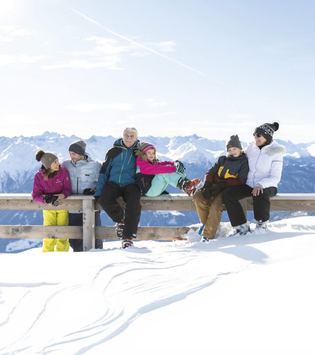 Eine Gruppe sitzt an einem Aussichtspunkt auf der Holzbank im Schnee und schaut ins Tal.