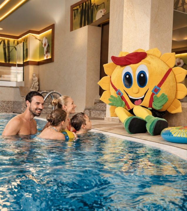 Eltern und zwei Kinder im Innenpool, daneben sitzt das Maskottchen Sonni am Beckenrand.