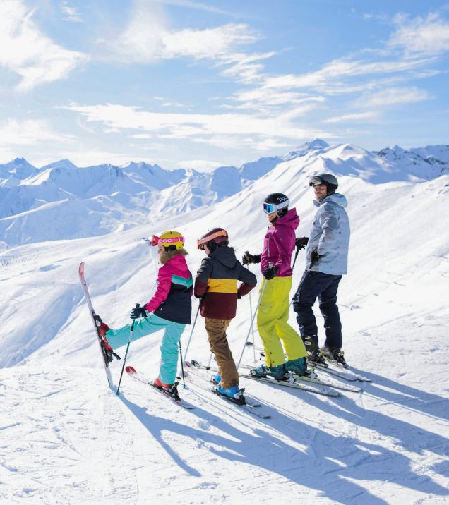 A family with skis at the edge of the piste in front of the snowy mountain panorama.