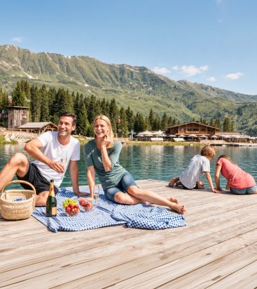 Ein Paar beim Picknick auf dem Steg am See bei Serfaus, Kinder spielen am Ufer, Berge im Hintergrund.