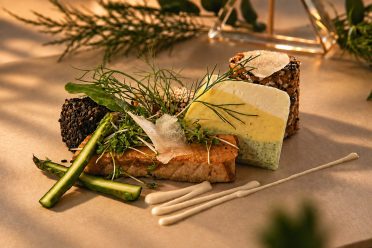 Fischfilet mit grünem Spargel, Kräutern und zweierlei Brot auf hellem Tisch.