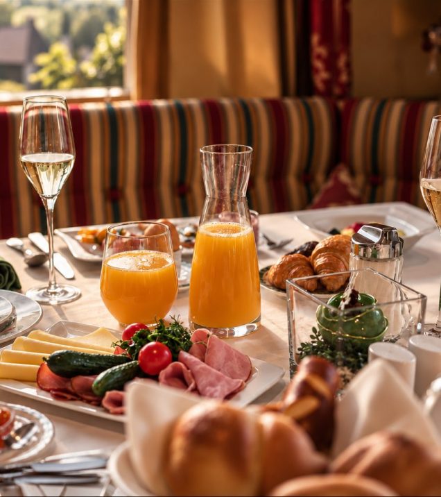Breakfast table laid with orange juice, sparkling wine, cheese, cold cuts and pastries in front of a window.