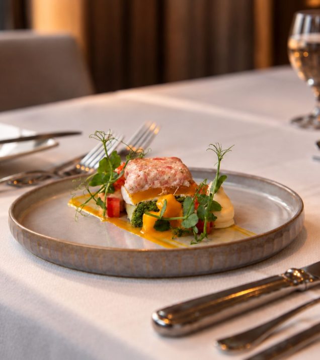 Fish course with vegetables on a plate, cutlery and a wine glass beside it.