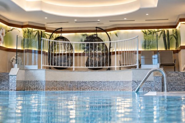 Innenpool im Hotel DreiSonnen, dahinter zwei Hängesessel hinter einem weißen Geländer und Liegen, im Vordergrund unscharf grüne Pflanzen.