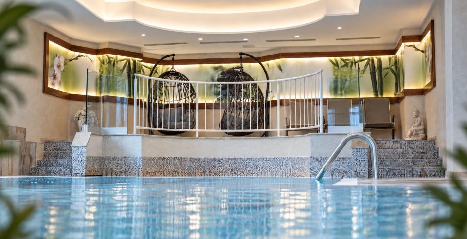 Innenpool im Hotel DreiSonnen, dahinter zwei Hängesessel hinter einem weißen Geländer und Liegen, im Vordergrund unscharf grüne Pflanzen.