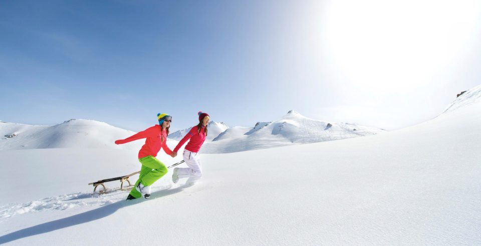 Zwei Kinder laufen mit ihrem Schlitten durch den tiefen Schnee, im Gegenlicht die weite Hochfläche.