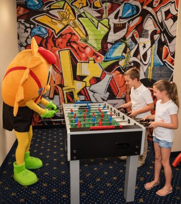 The mascot Sonni playing table football with two children in front of the graffiti wall at Hotel DreiSonnen in Serfaus