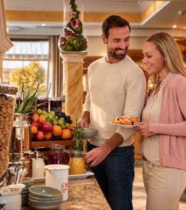 Ein Paar steht am Frühstücksbuffet und stellt sich einen Teller mit Gebäck zusammen, daneben Müslispender und Obst.