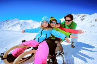 Eine Familie sitzt zu viert auf zwei Schlitten im Schnee und breitet lachend die Arme aus.