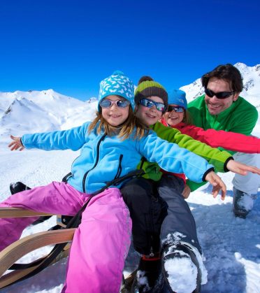 Eine Familie sitzt zu viert auf zwei Schlitten im Schnee und breitet lachend die Arme aus.