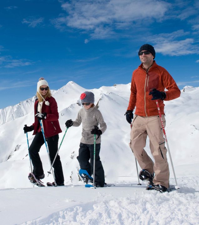 Eine Familie geht mit Schneeschuhen und Stöcken durch den Schnee, dahinter die weiße Bergkette.