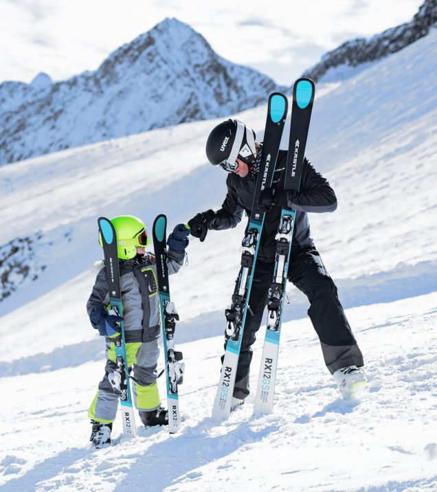 Adult and child with their skis upright in the snow in front of the mountain panorama.