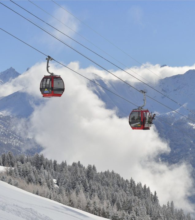 Zwei Gondeln schweben über dem verschneiten Wald, dahinter die Berge und ziehende Wolken.