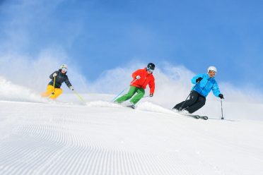 Three adults carve side by side down the freshly groomed piste.