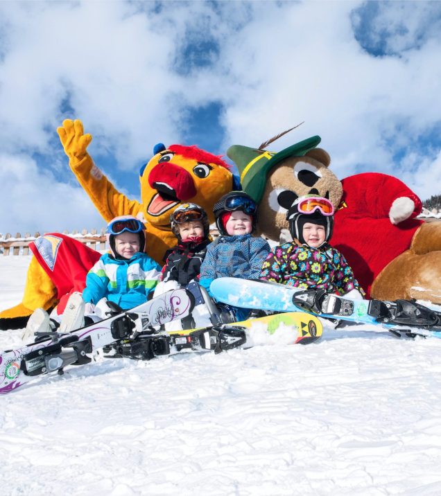 Kinder liegen mit ihren Snowboards im Schnee, dahinter zwei Maskottchen in bunten Kostümen.