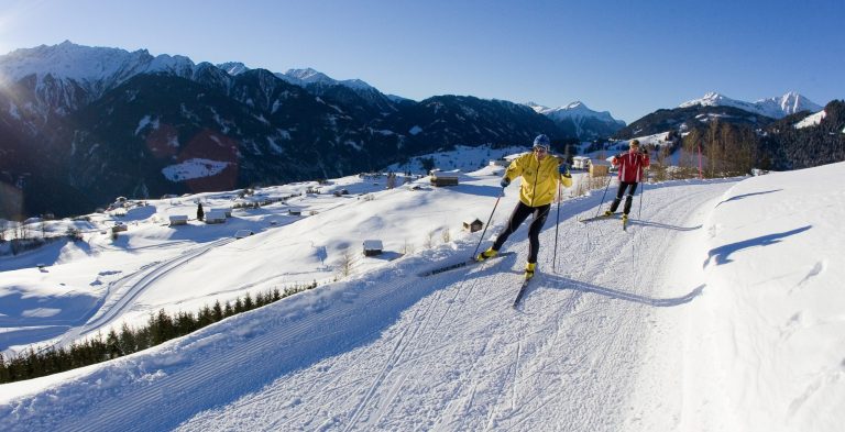 Two cross-country skiers on the groomed trail high above the snowy village, the mountains behind.
