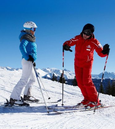 Eine Skifahrerin und ein Skilehrer der Skischule Serfaus stehen auf der sonnigen Piste über dem Tal.