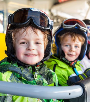 Zwei Kinder mit Helm und Skibrille schauen lachend in die Kamera.