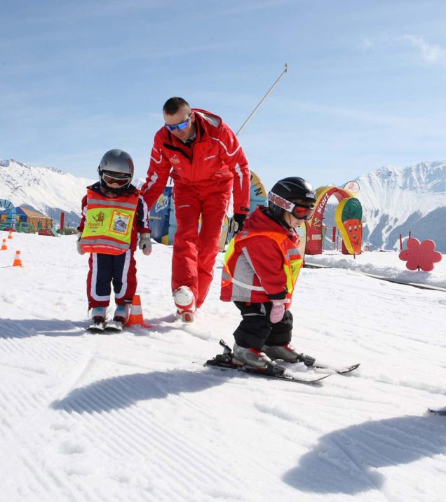Ein Skilehrer führt zwei Kinder über den flachen Übungshang, daneben die bunten Tore des Kinderareals.