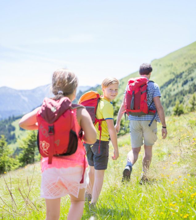 A family hiking across a summer meadow above Serfaus, three people with rucksacks seen from behind.