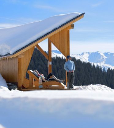 Two people at a wooden shelter in the snow, one lying on the panorama lounger, behind them the snow-covered mountains.