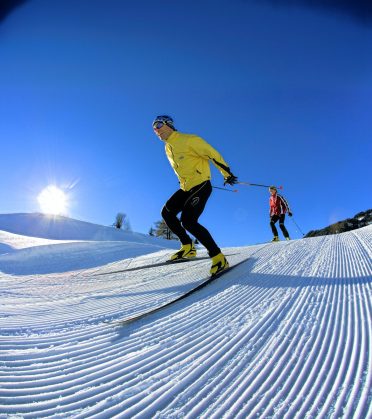 A skier glides over the freshly groomed piste, behind him a second person and the low sun.