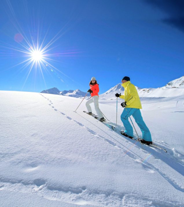 Two people on skis make their tracks through untouched snow, the sun above them in a deep blue sky.
