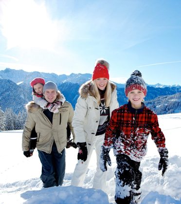 Eine Familie geht nebeneinander über den verschneiten Weg, dahinter die Gipfelkette.