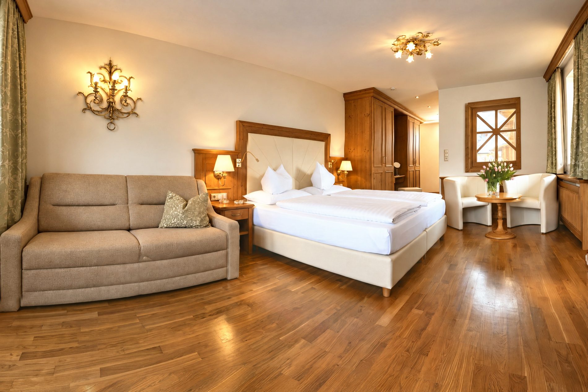 Doppelbett mit gepolstertem Kopfteil, Sofa und Sitzecke am Fenster in einer vergleichbaren Deluxe-Suite vom Typ des Zimmers Elias im Hotel DreiSonnen in Serfaus