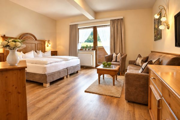 Double bed with carved headboard, seating group and balcony door looking out on greenery in a comparable room of the Suite Elisabeth type at Hotel DreiSonnen in Serfaus