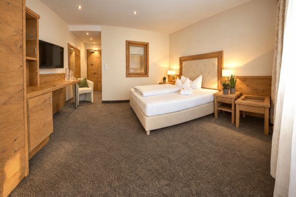 Bed with upholstered headboard, desk with TV and built-in oak wardrobe in the Florian room at Hotel DreiSonnen in Serfaus