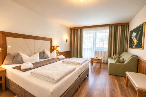 Double bed with upholstered headboard, green sofa and a wall of windows with mountain views in the Comfort Double Room Jonas at Hotel DreiSonnen in Serfaus