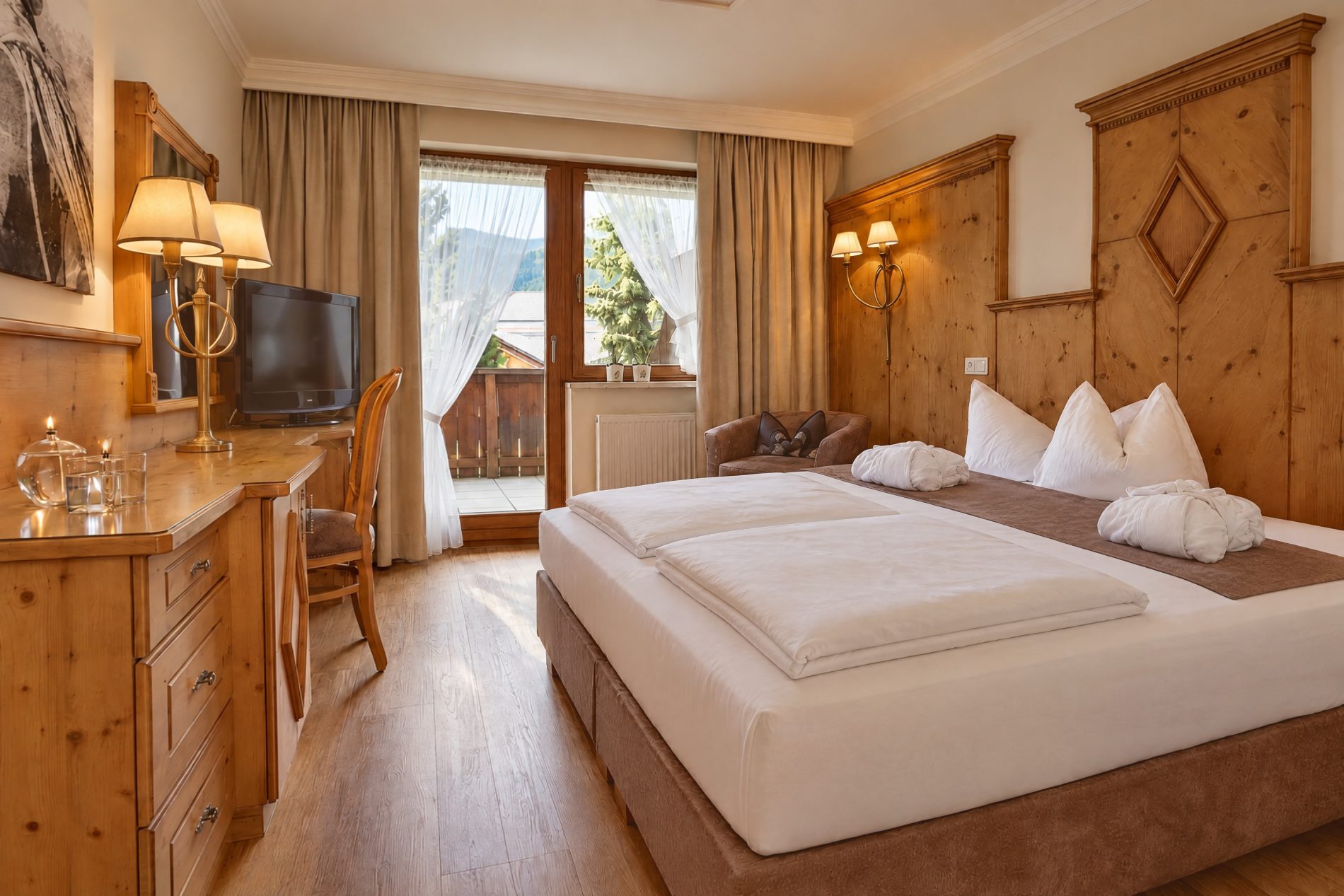 Doppelbett vor der Holzvertäfelung mit Rautenmotiv, Schreibtisch und Balkontür im Komfort-Einzelzimmer König Ludwig des Hotels DreiSonnen in Serfaus