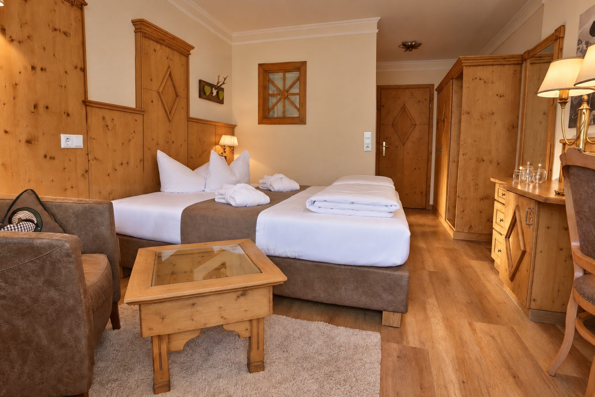 Doppelbett, Couchtisch mit Hirschfigur und Blumenstrauß und Kleiderschrank aus Holz im Zimmer König Ludwig des Hotels DreiSonnen in Serfaus