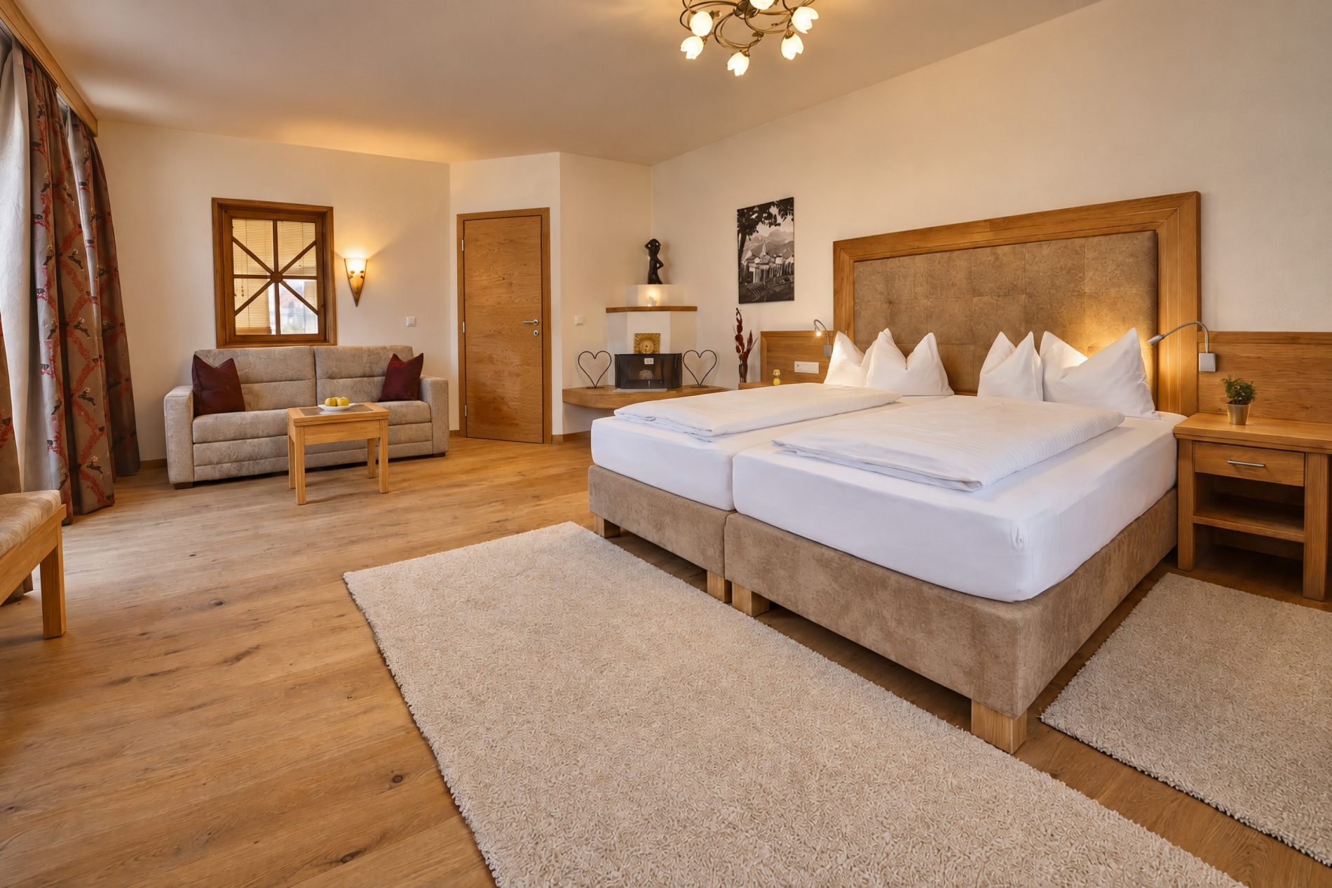 Doppelbett mit gepolstertem Kopfteil, Teppich und Sofaecke unter dem Sprossenfenster in der Suite Magdalena des Hotels DreiSonnen in Serfaus