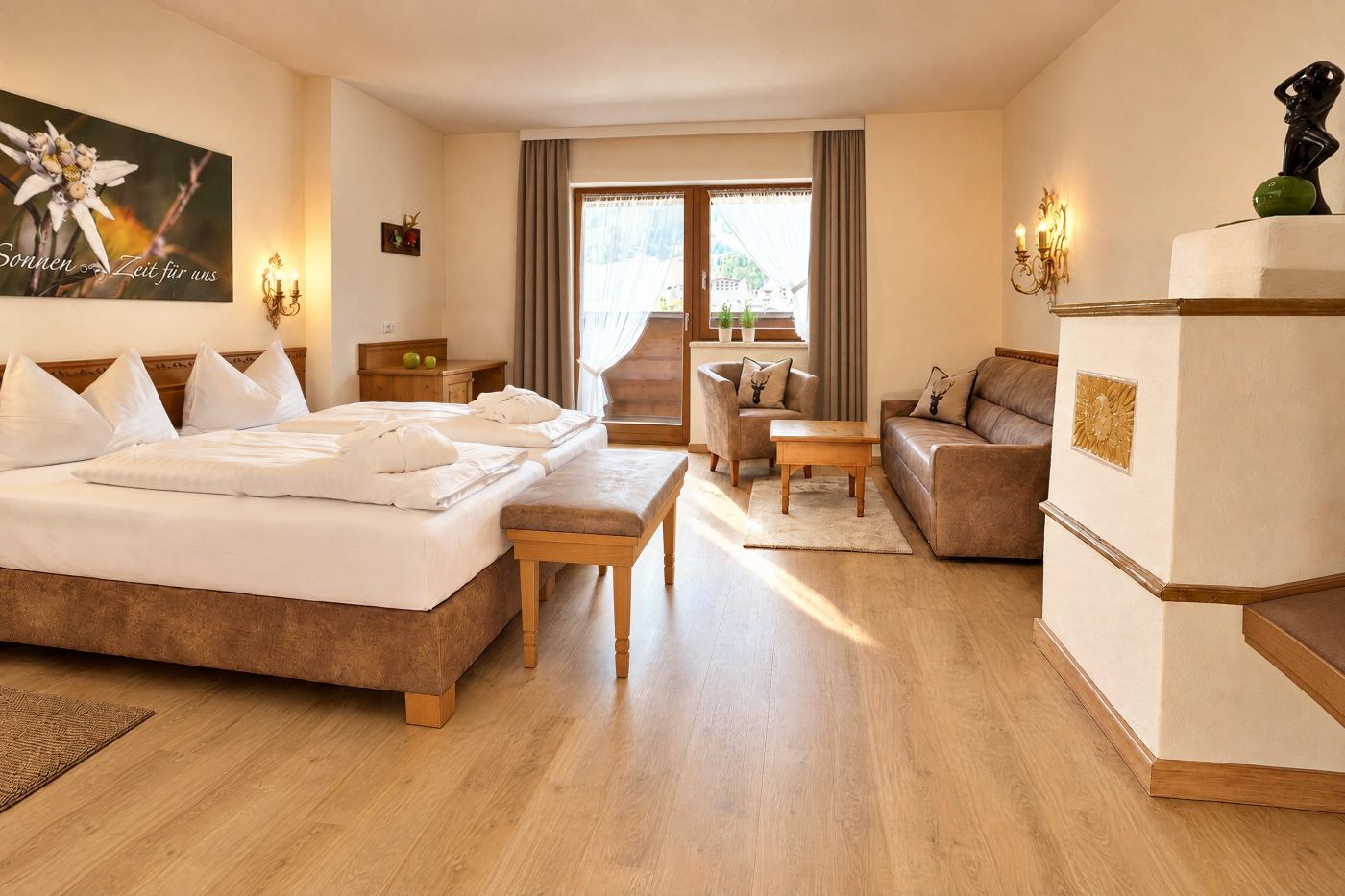 Double bed, seating group in front of the balcony door and tiled stove in the Valentina room at Hotel DreiSonnen in Serfaus