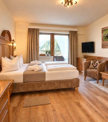 Doppelzimmer Viktoria im Hotel DreiSonnen in Serfaus mit geschnitztem Holzbett, Sitzecke und Balkontür mit Blick in den Wald
