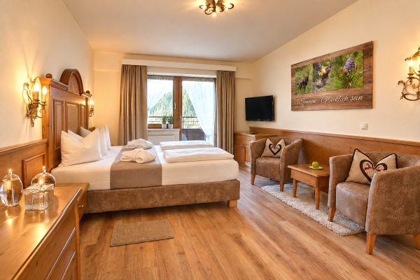 Double bed with a carved headboard, two armchairs with a sparkling wine cooler and balcony door in the Double Room Viktoria at Hotel DreiSonnen in Serfaus