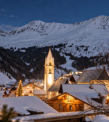 Verschneites Bergdorf mit Kirchturm und erleuchteten Häusern am Abend