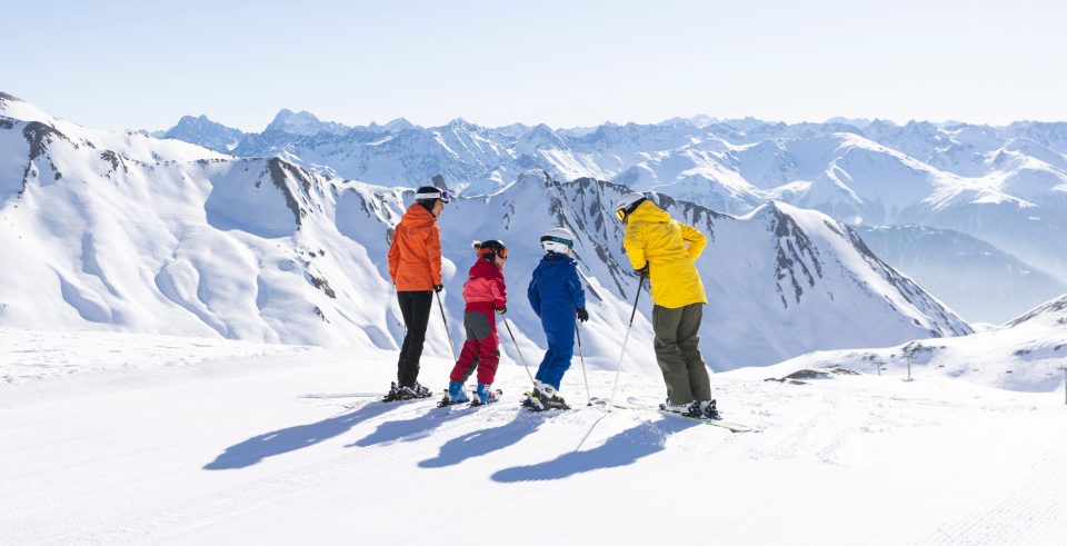 Vier Menschen in bunten Skijacken auf der Piste vor verschneiten Gipfeln