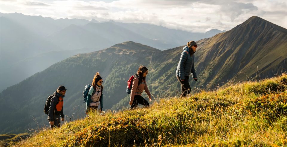Vier Wandernde steigen einen grasigen Hang hinauf, dahinter Bergketten im Gegenlicht