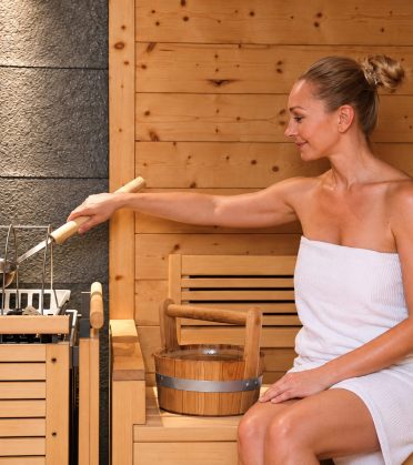 Frau im Handtuch gießt in der Sauna Wasser über die heißen Steine