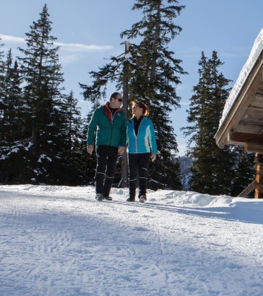 Paar beim Winterspaziergang zwischen verschneiten Fichten neben einer Holzhütte