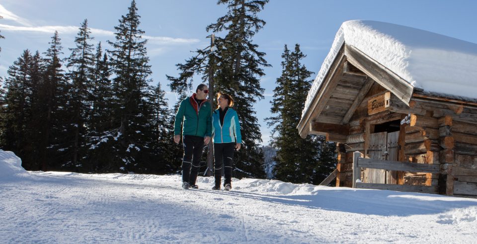 Paar beim Winterspaziergang zwischen verschneiten Fichten neben einer Holzhütte