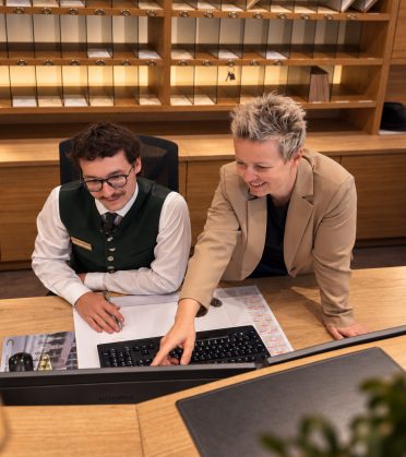 At the reception of the Hotel DreiSonnen, a colleague shows a young employee something on the keyboard, both smiling.